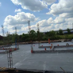 Wylana płyta fundamentowa z wystającymi pionowymi zbrojeniami i rurami instalacyjnymi na tle błękitnego nieba z obłokami.