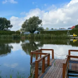 Spokojny krajobraz z drewnianym pomostem nad stawem, odbicie drzew w wodzie, w tle żywopłot i żółta ciężarówka.