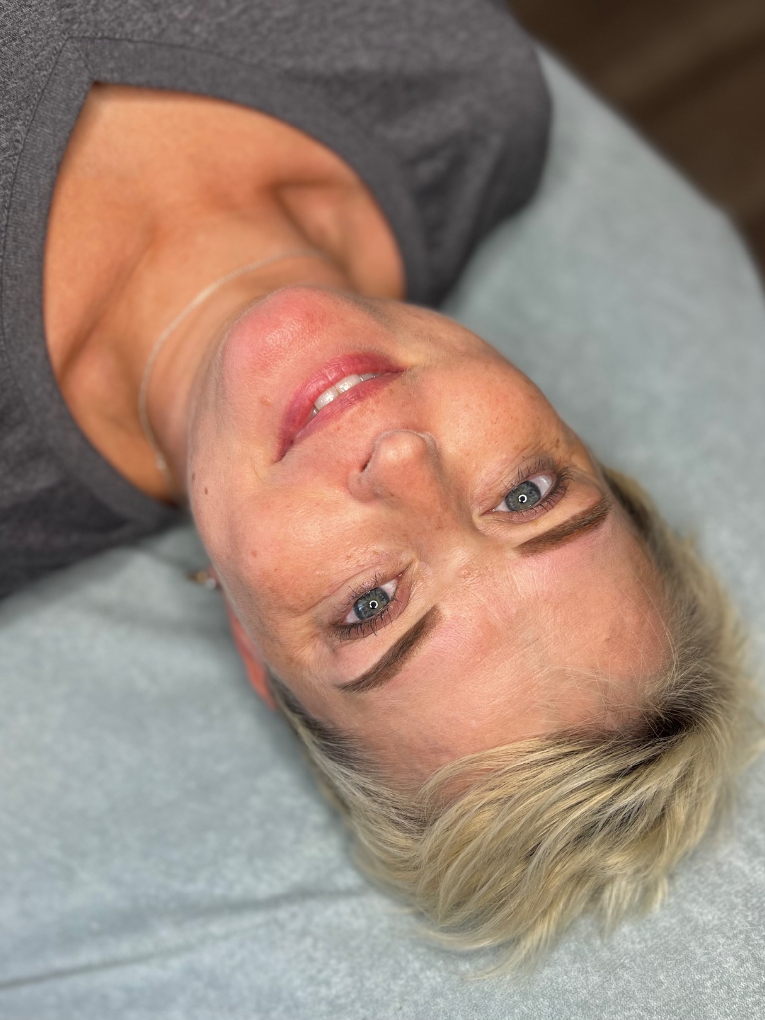 Kobieta z blond włosami leżąca na jasnym podłożu, prezentująca efekt makijażu permanentnego brwi metodą ombre, z widocznymi delikatnymi przejściami kolorystycznymi i naturalnym wyglądem.