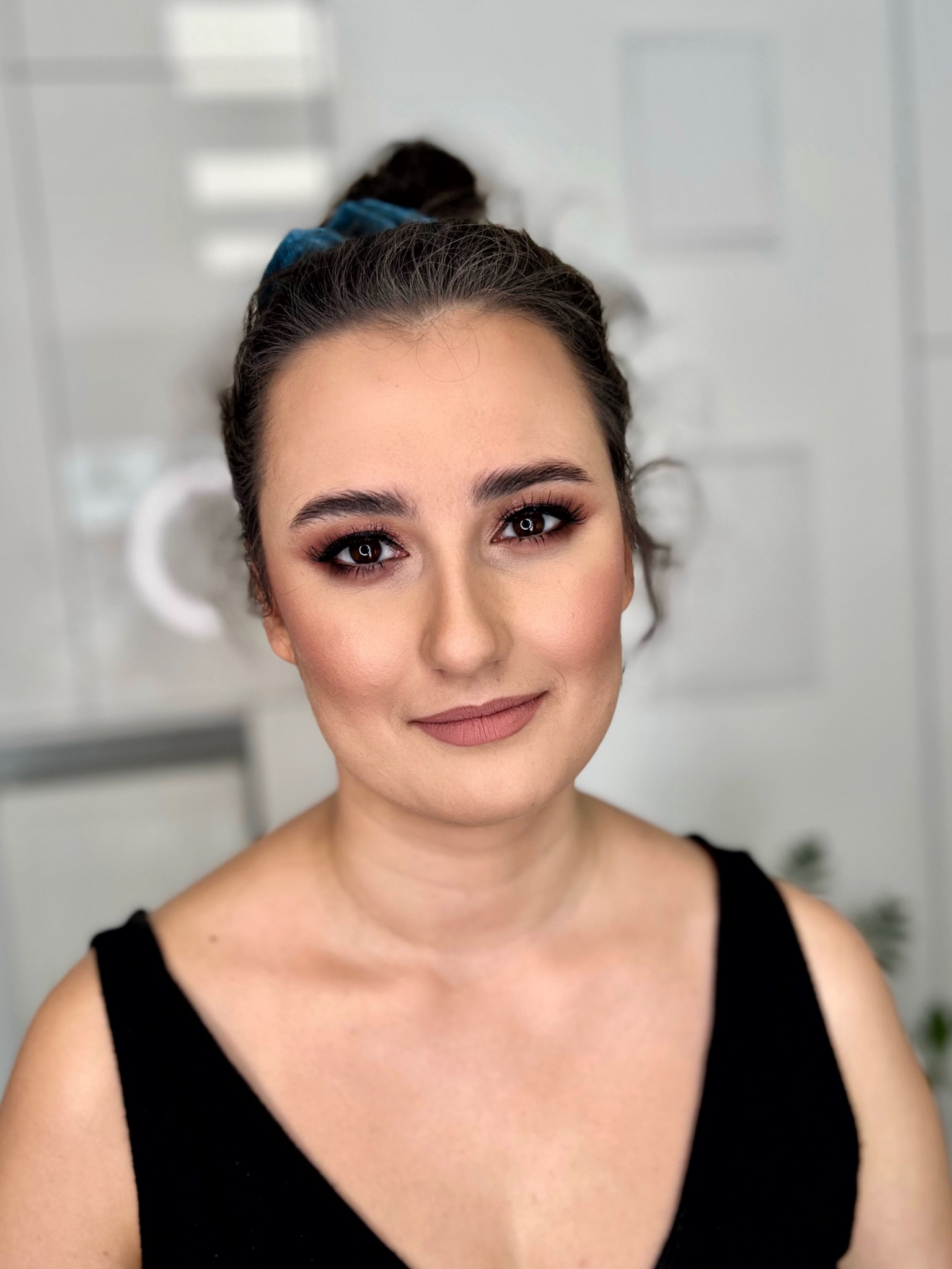 Portret kobiety z profesjonalnym makijażem, uwydatnione oczy i usta w odcieniach brązu, włosy upięte w kok z niebieską gumką, w tle rozmyte elementy salonu.