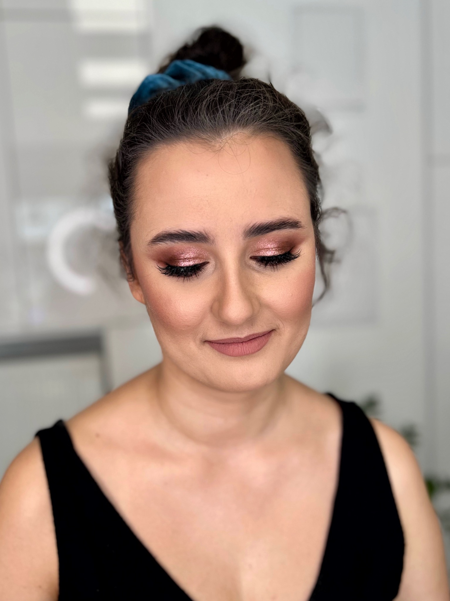 Portret kobiety z zamkniętymi oczami, prezentującej makijaż oczu w odcieniach różowego złota i matową pomadkę w kolorze nude, włosy upięte w kok z niebieską gumką.