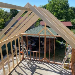 Konstrukcja drewniana altany w trakcie budowy, widoczne krokwie dachu i szkielet ścian, na tle zieleni ogrodowej i sąsiednich domów.