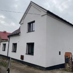 Dwukondygnacyjny dom z jasną elewacją, ciemnymi oknami i dachem z brązową obróbką, widoczny z boku, z metalowym ogrodzeniem i skrzynką pocztową z czerwonym daszkiem.