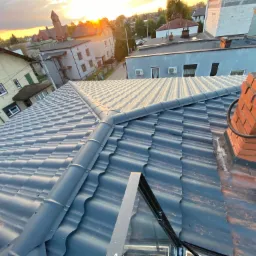 Nowy, grafitowy dach z blachodachówki modułowej, z obróbką blacharską i kominem z cegły na tle zabudowy miejskiej o zachodzie słońca.