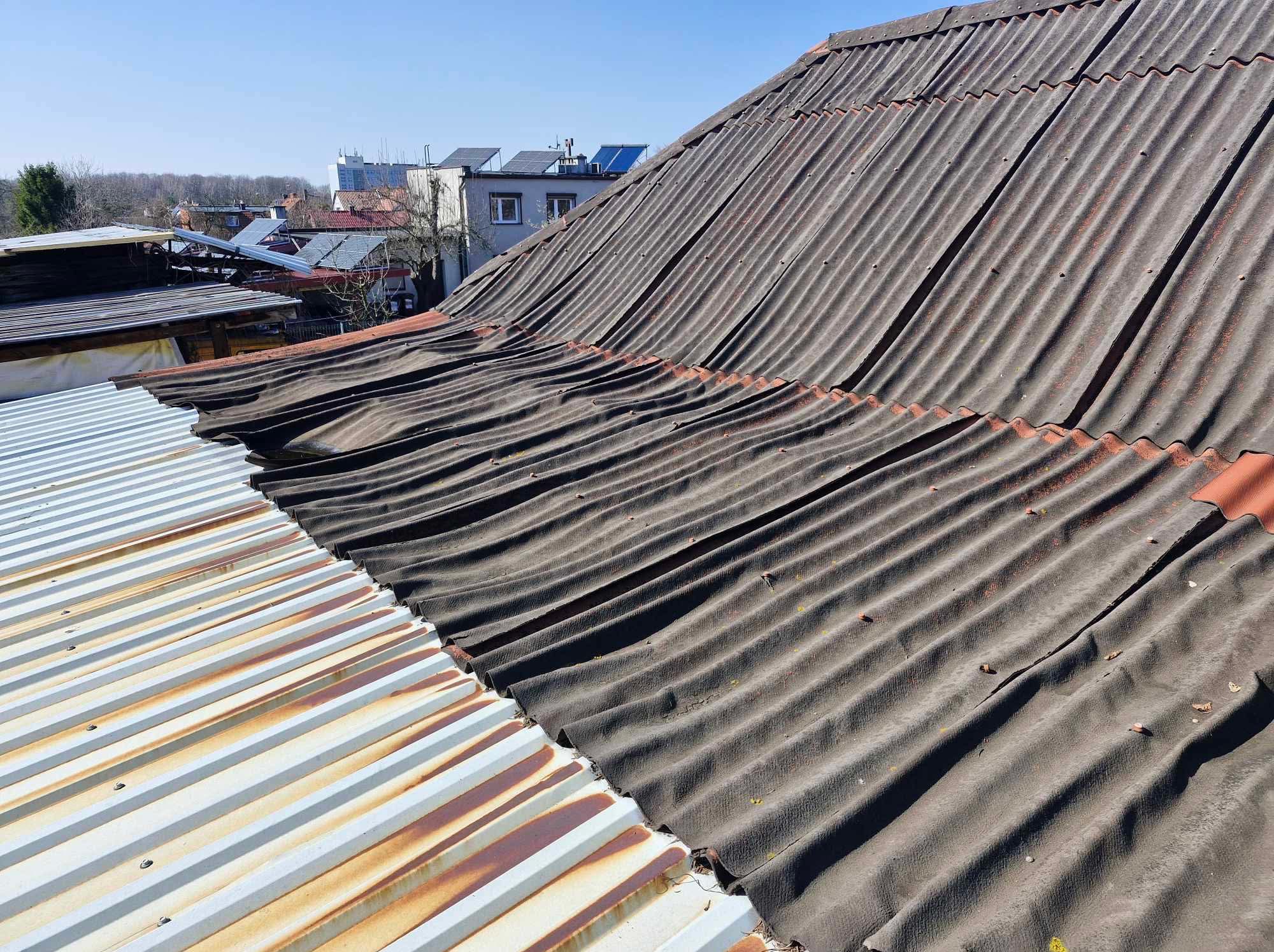Falisty dach z eternitu w odcieniach szarości, z widocznymi śladami zużycia i zabrudzeń, sąsiadujący z fragmentem dachu z blachy trapezowej, zardzewiałej w niektórych miejscach.