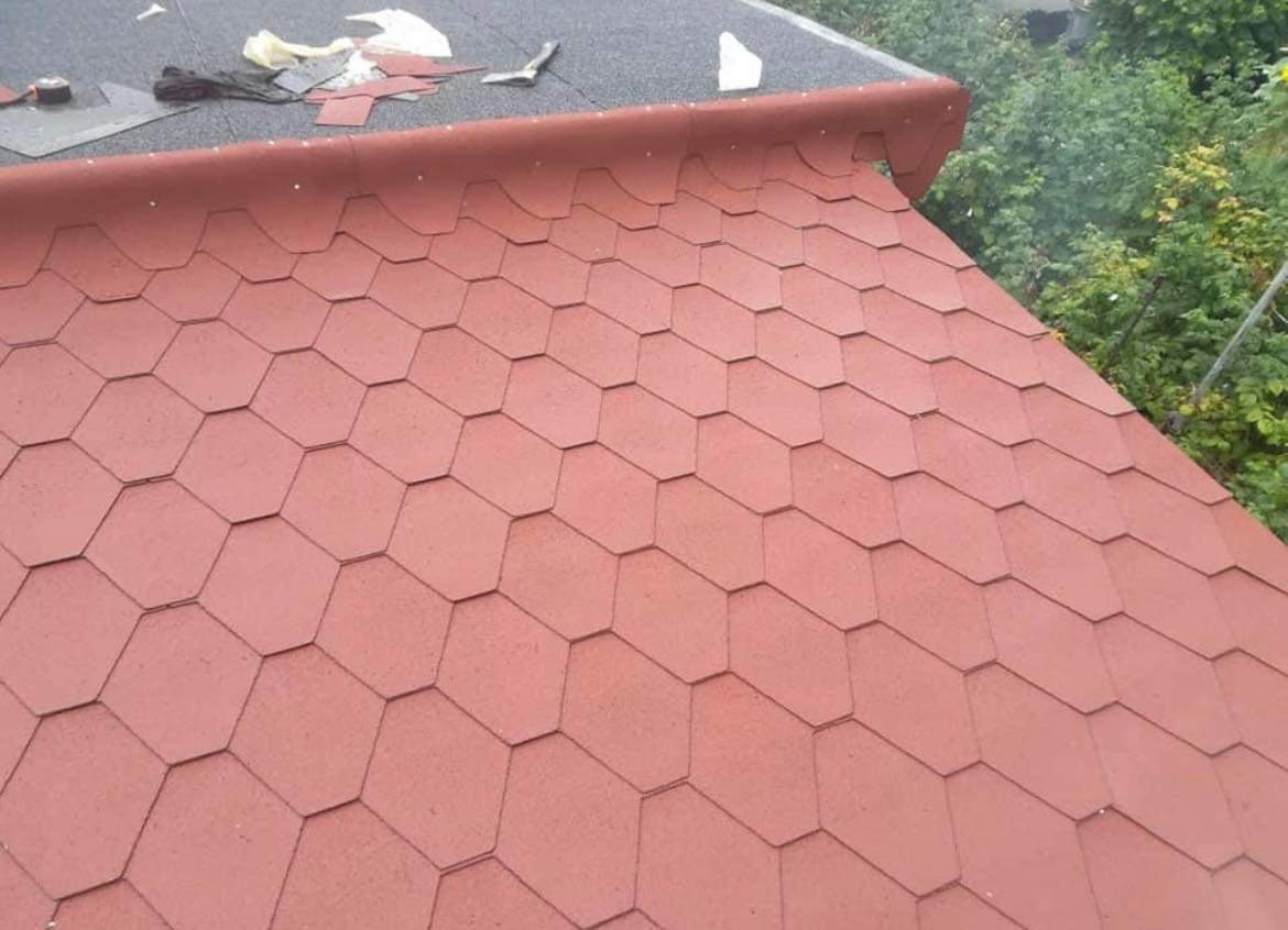 Układanie czerwonej dachówki bitumicznej na dachu, widoczne narzędzia i resztki materiałów na czarnej powierzchni dachu w tle.