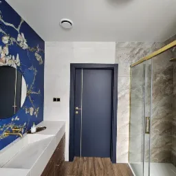 Nowoczesna łazienka z akcentem artystycznym: ściana z motywem kwitnących gałęzi migdałowca Van Gogha, okrągłe lustro, minimalistyczna umywalka z drewnianą szafką, kabina prysznicowa z złotymi...