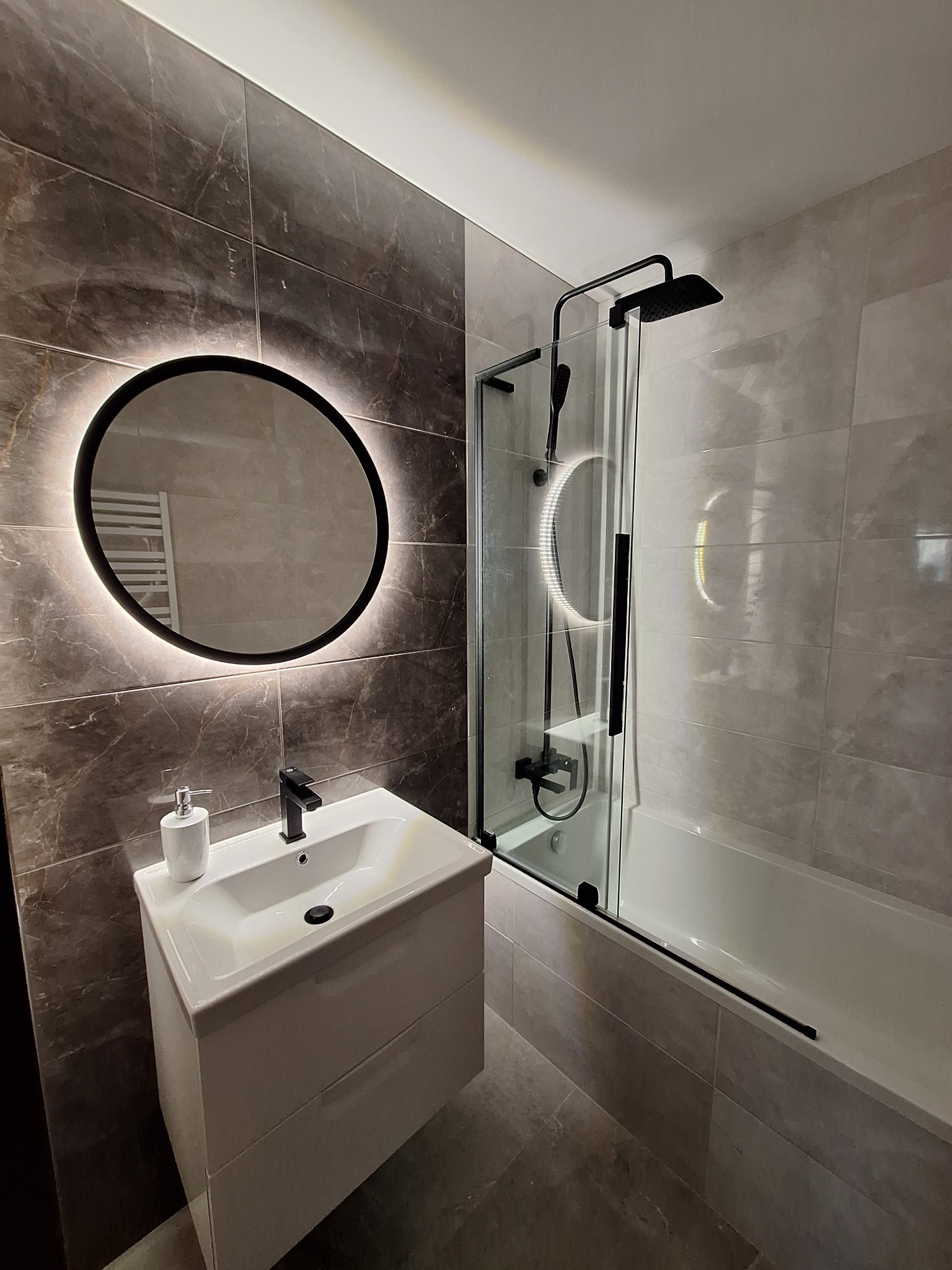 Nowoczesna łazienka z marmurowymi płytkami, okrągłym lustrem z podświetleniem LED, białą umywalką nablatową i czarną armaturą oraz kabiną prysznicową z czarnymi elementami.