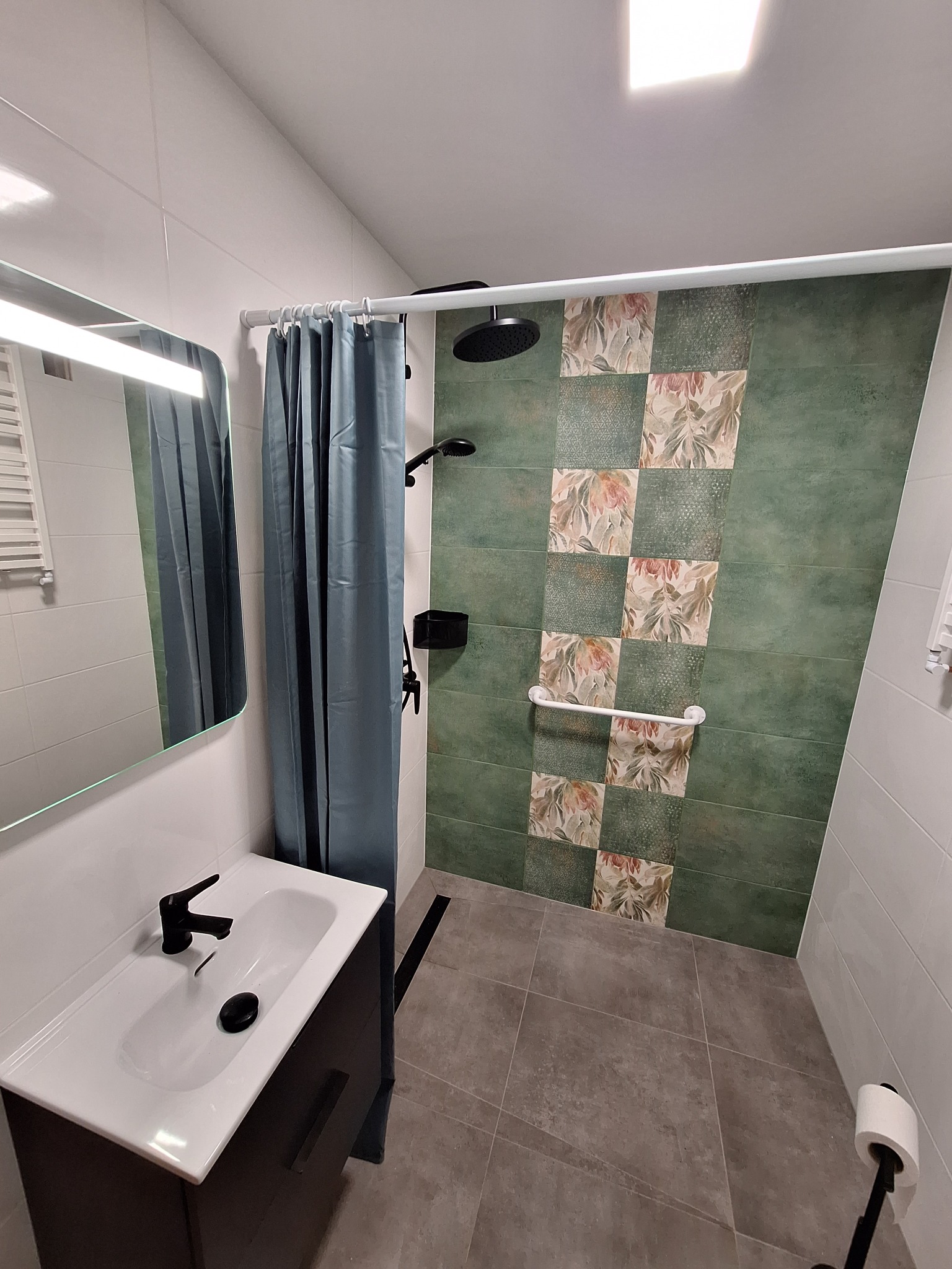 Nowoczesna łazienka z zielonymi płytkami w kwiatowe wzory, czarną armaturą i zasłoną prysznicową w kolorze niebieskim.
