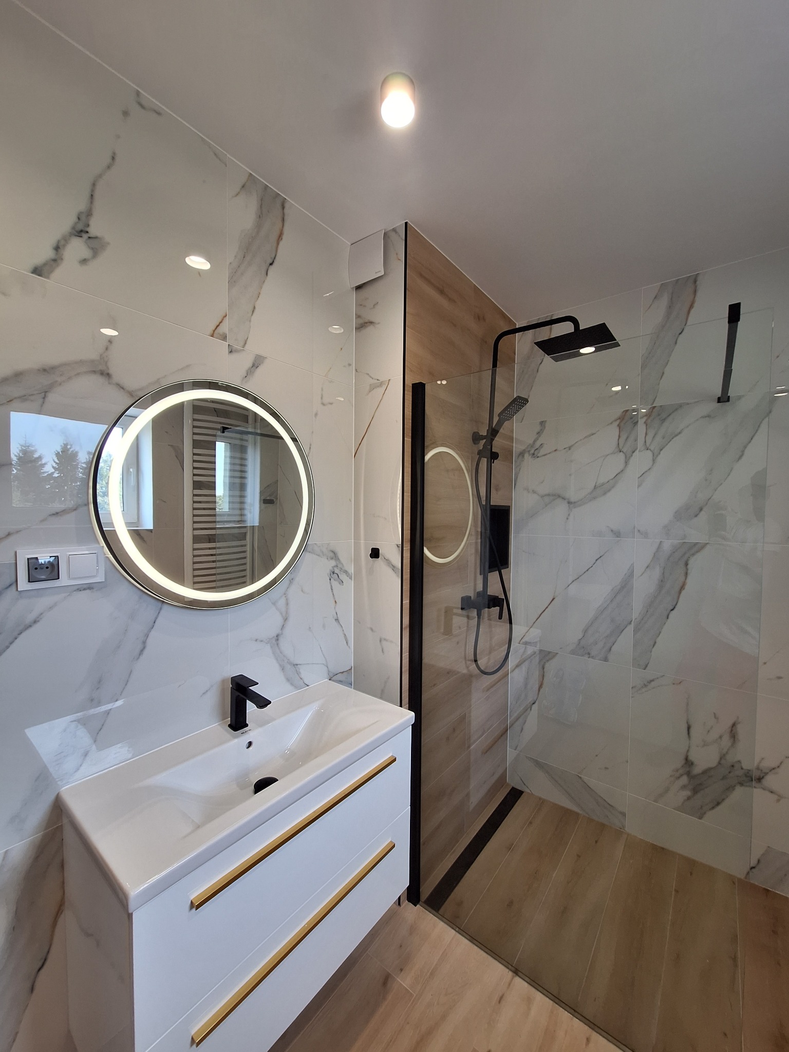 Nowoczesna łazienka z kabiną prysznicową typu walk-in, umywalką nablatową z czarną armaturą i okrągłym lustrem z podświetleniem LED, wykończona płytkami imitującymi marmur i drewnem.