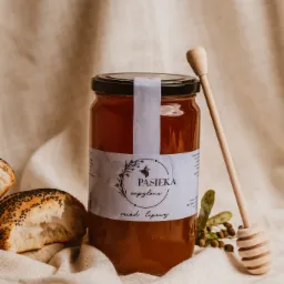 Słoik miodu lipowego z etykietą 'Pasieka zapylone' i drewnianym nabierakiem, obok kawałek bułki z makiem na tle beżowej tkaniny.