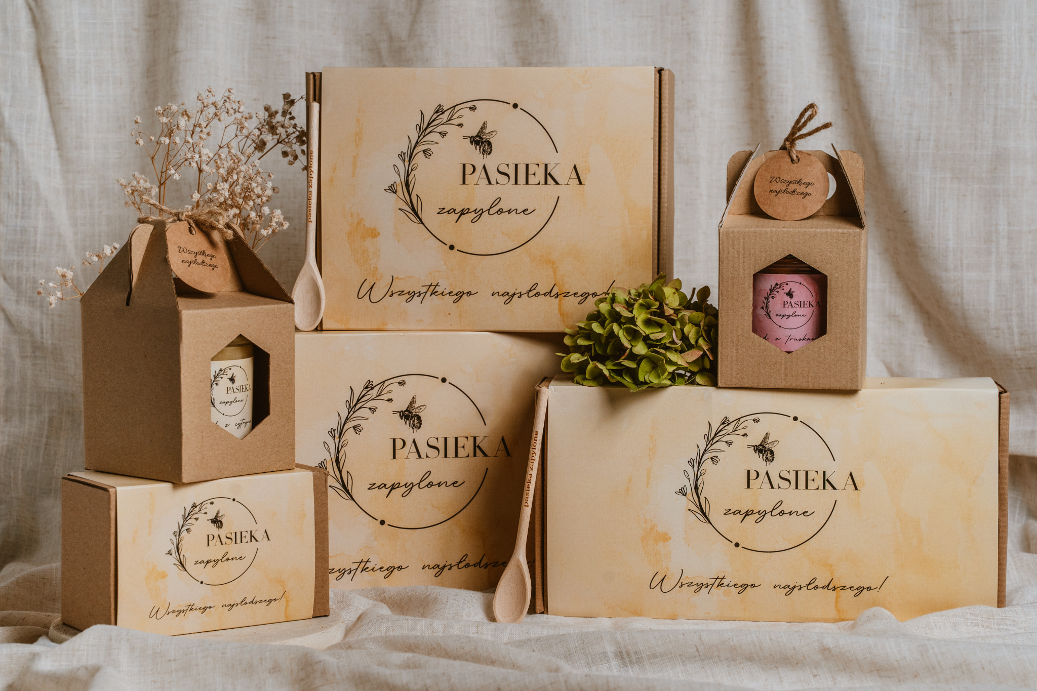 Zestaw prezentowy w beżowych pudełkach z logo 'Pasieka zapylone' i napisem 'Wszystkiego najsłodszego!', drewniana łyżeczka i suszone kwiaty na kremowym tle.