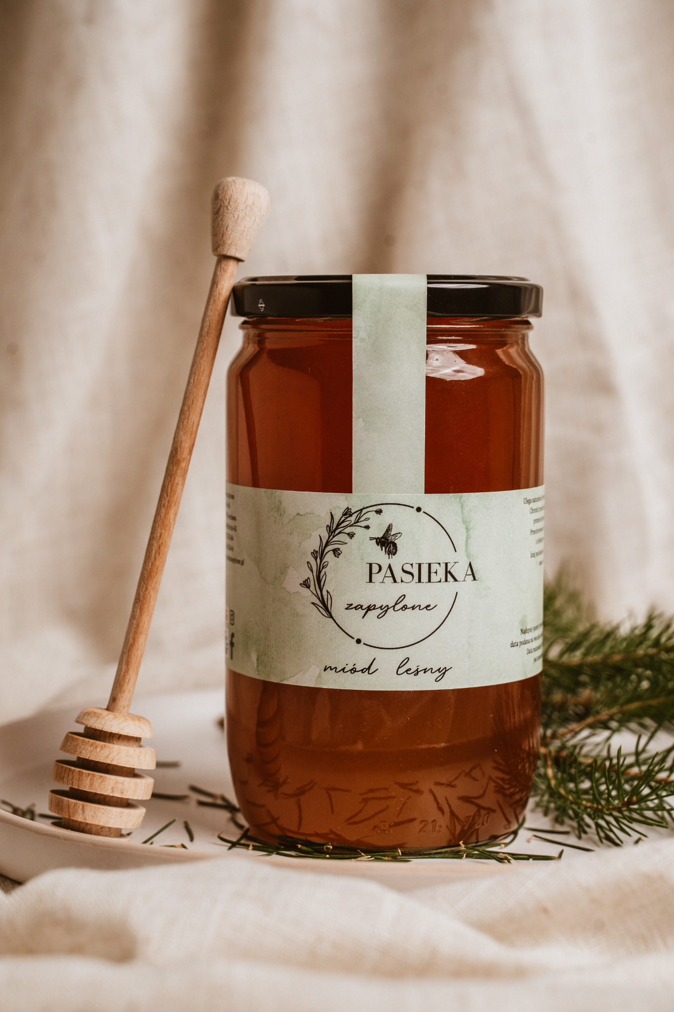 Słoik miodu leśnego z drewnianym nabierakiem na tle kremowego lnu i gałązki świerku, etykieta z napisem 'Pasieka zapylone'.