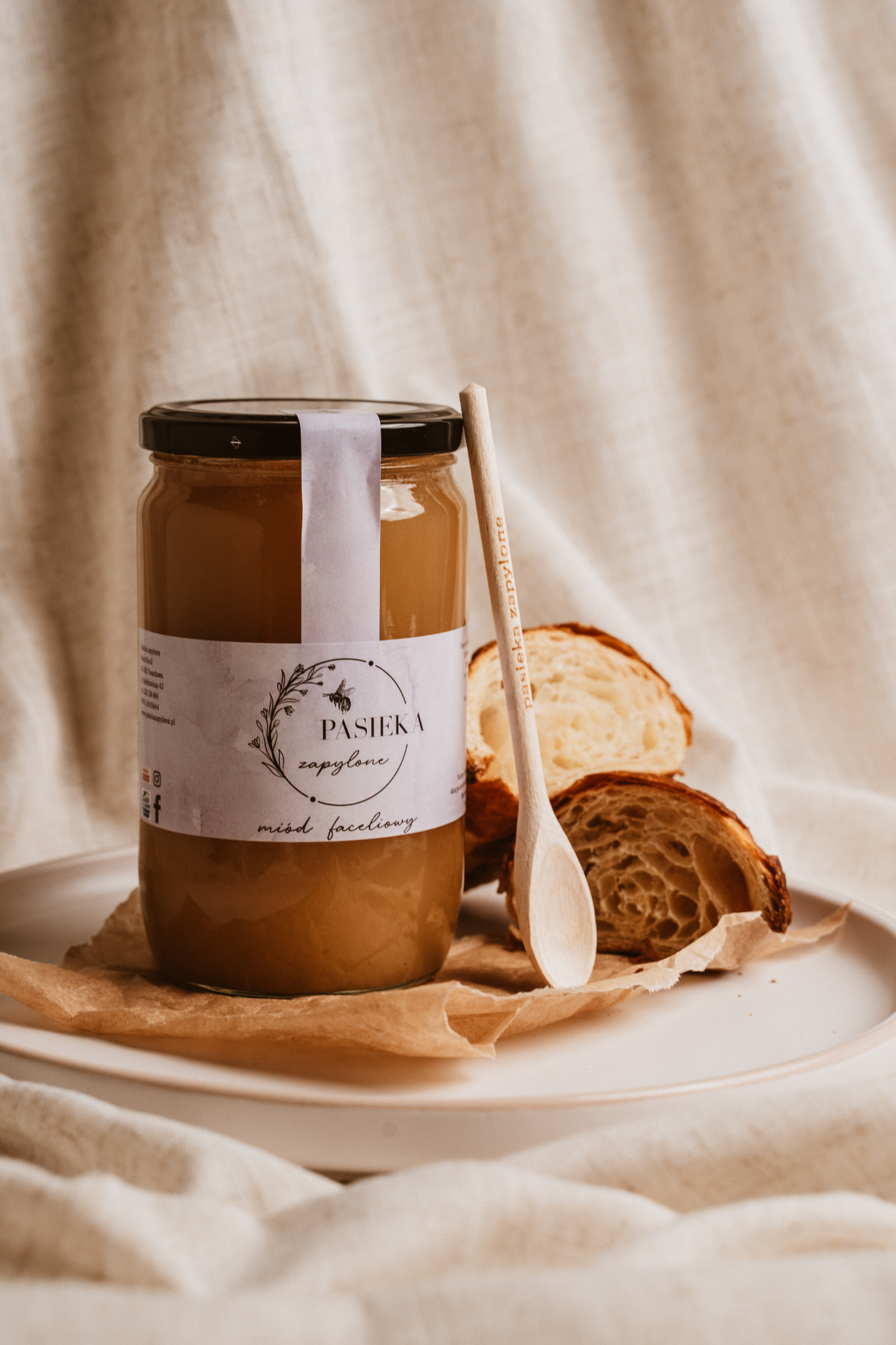 Słoik miodu faceliowego z etykietą Pasieka Zapylone, drewniana łyżka z nadrukiem nazwy pasieki i dwa kawałki croissanta na białym talerzu z beżowym papierem w tle, na tle beżowej tkaniny.