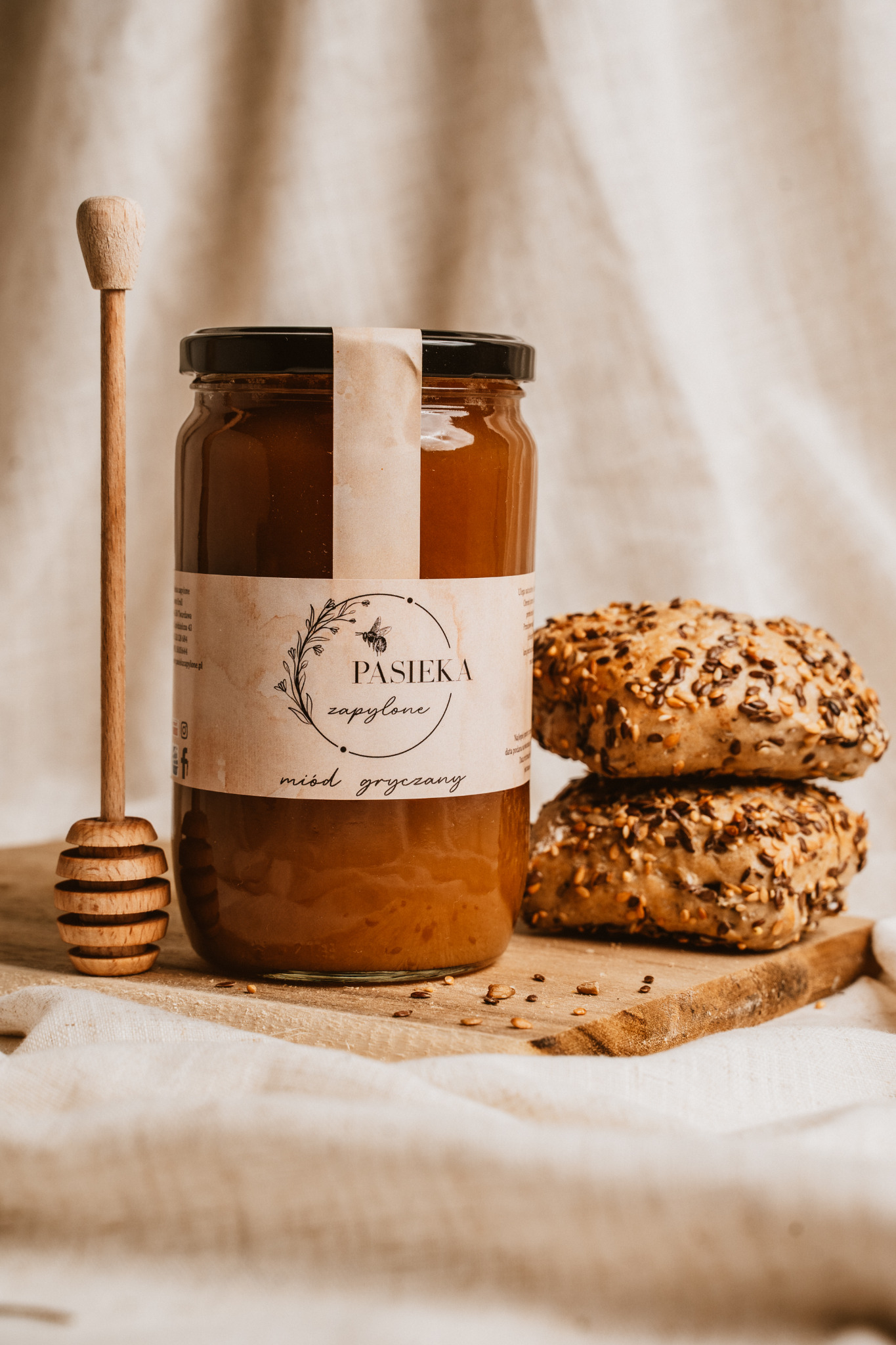 Słoik miodu gryczanego z drewnianym nabierakiem i dwoma bułeczkami z ziarnami na drewnianej desce, całość na lnianym tle.