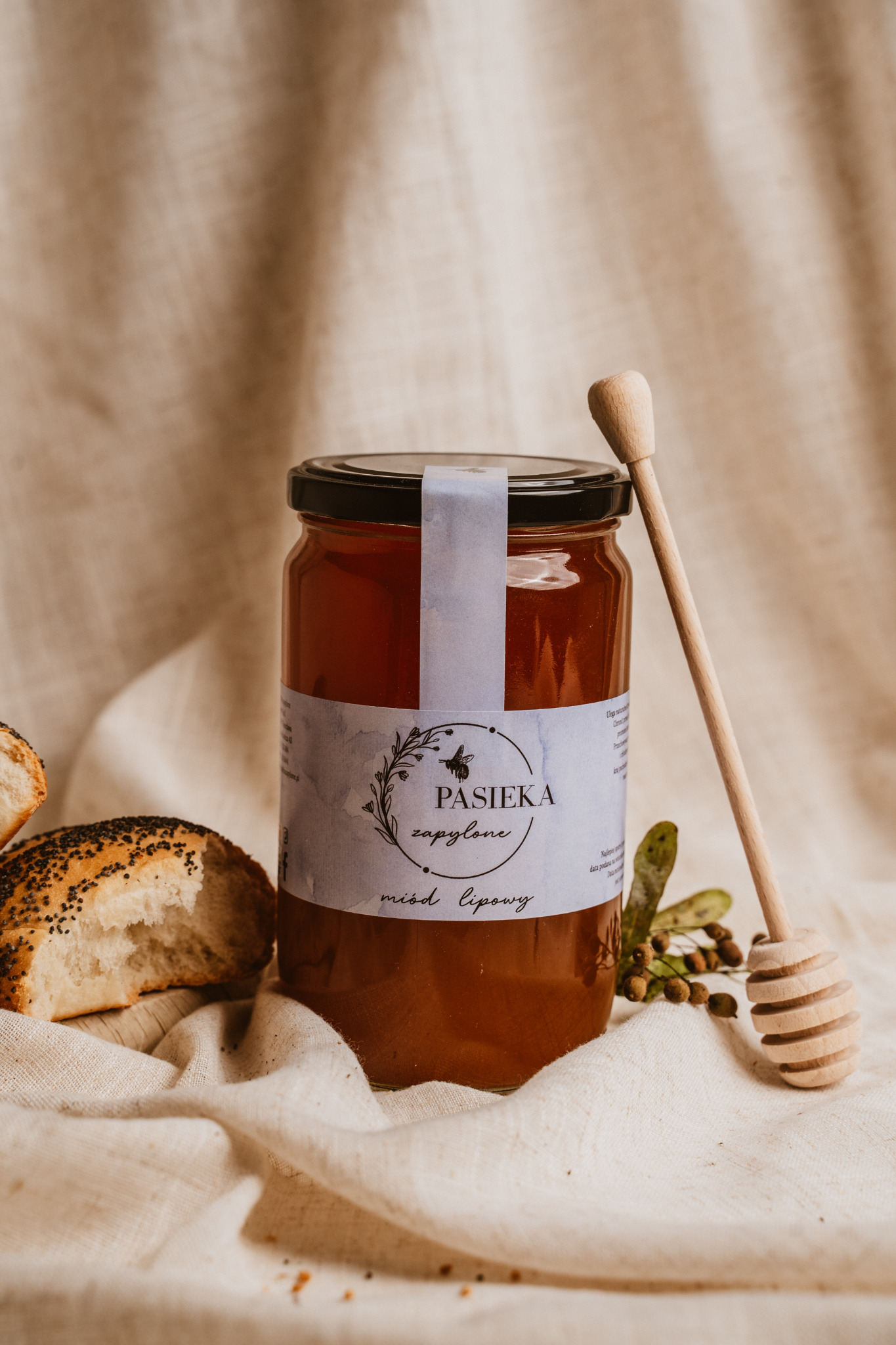 Słoik miodu lipowego z etykietą 'Pasieka zapylone' i drewnianym nabierakiem, obok kawałek bułki z makiem na tle beżowej tkaniny.
