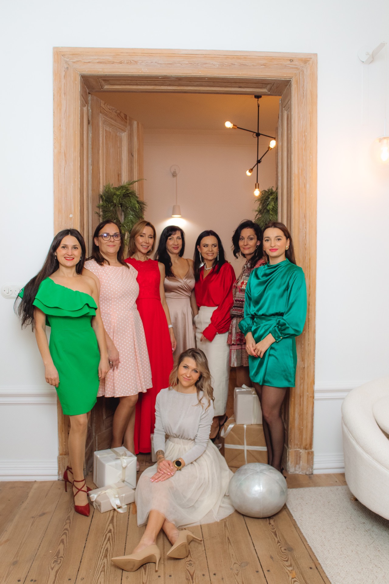 Grupa kobiet, prawdopodobnie pośredniczek, w eleganckich sukienkach, pozuje w drzwiach w jasnym wnętrzu z prezentami, sugerując udane transakcje.