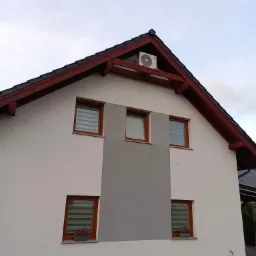 Biały dom jednorodzinny z brązowym dachem i oknami z roletami; na szczycie dachu zamontowana jednostka zewnętrzna klimatyzacji.