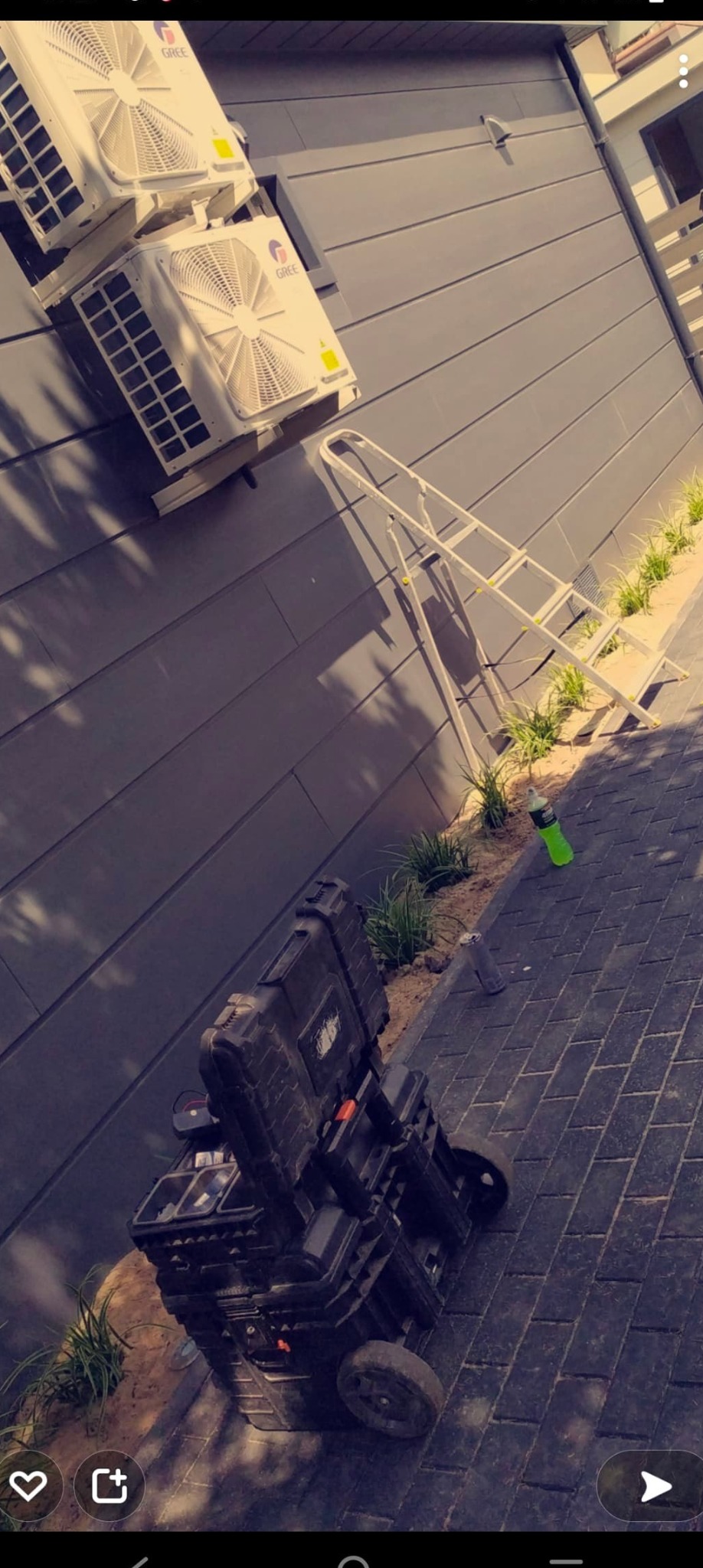 Dwa klimatyzatory marki Gree zamontowane na szarej elewacji budynku, obok drabina i skrzynka narzędziowa na kółkach oraz butelka po napoju.