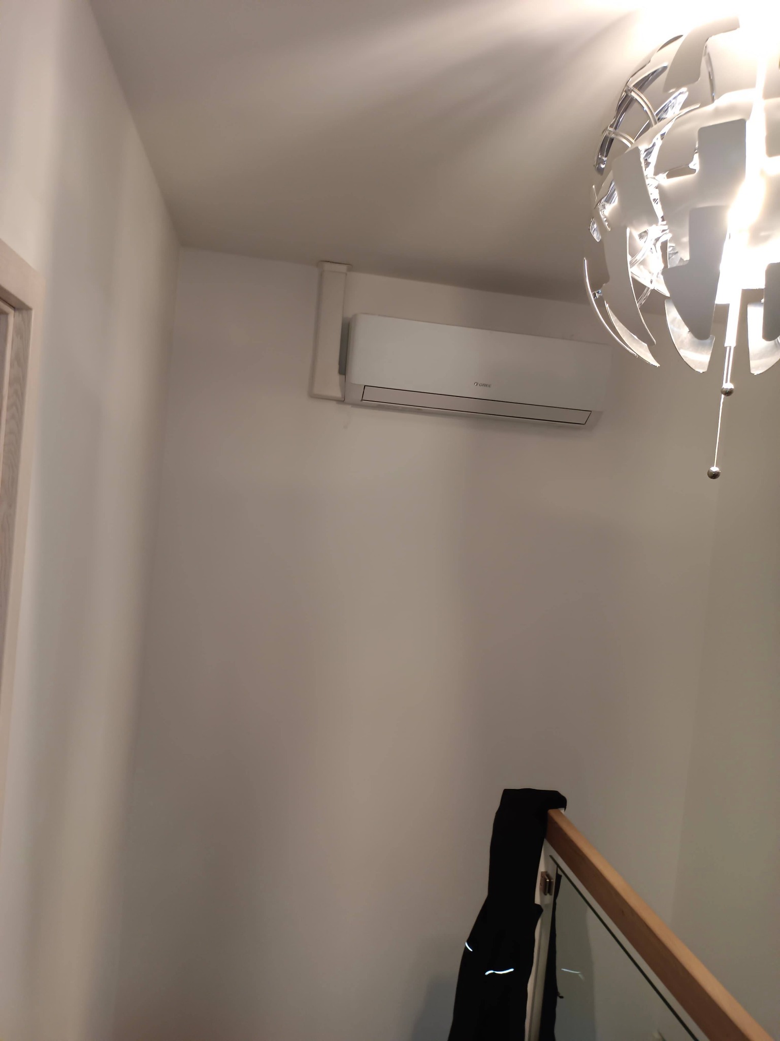 Biała klimatyzacja marki Gree zamontowana na ścianie w jasnym pomieszczeniu, widoczna z dołu, z dekoracyjną lampą sufitową i fragmentem balustrady schodów.