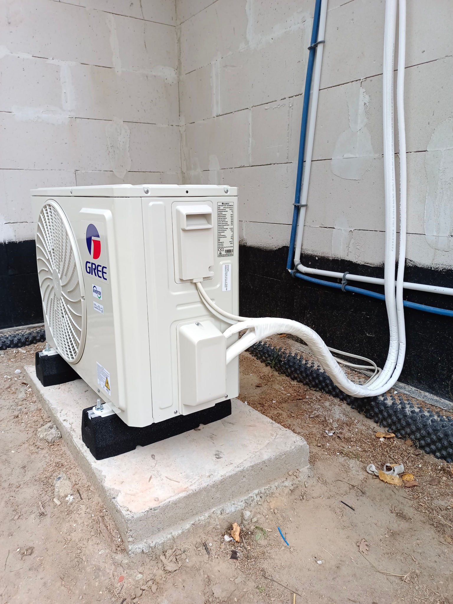 Zewnętrzna jednostka klimatyzacji Gree umieszczona na betonowym fundamencie, z widocznymi przewodami w izolacji, na tle ściany z bloczków betonowych i rur instalacyjnych.