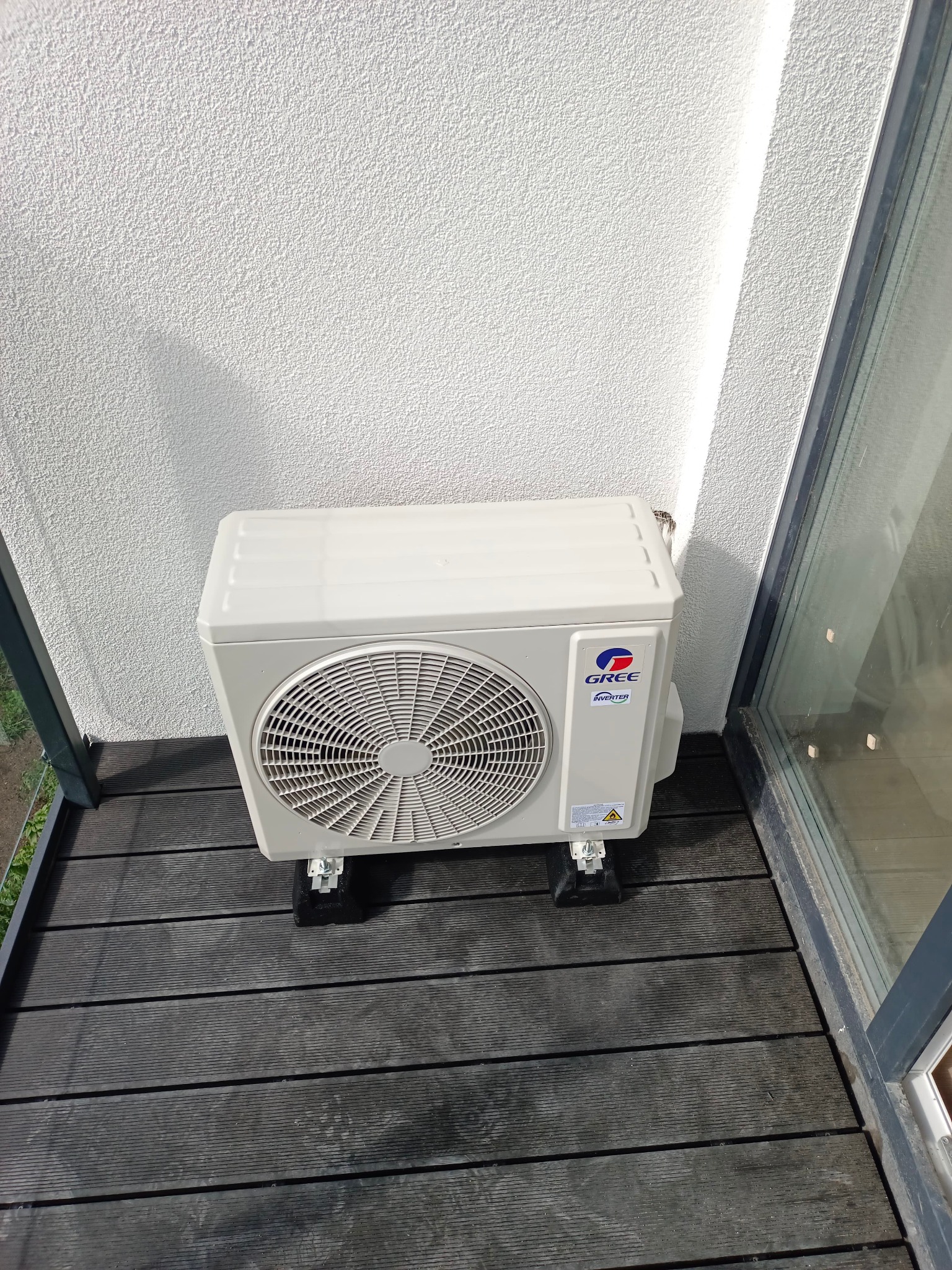 Zewnętrzna jednostka klimatyzacji marki Gree Inverter umieszczona na czarnych podkładkach na drewnianym balkonie, widoczna ściana budynku i fragment okna.