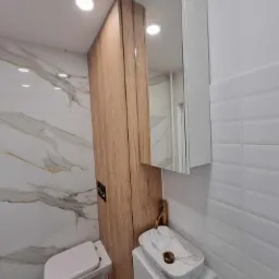 Nowoczesna łazienka z marmurowymi płytkami, drewnianą zabudową nad toaletą, białą umywalką nablatową z marmurowym wzorem i złotą armaturą oraz białymi, wypukłymi płytkami na ścianie.