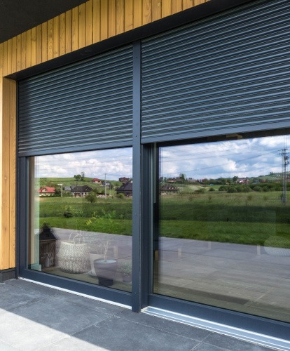 Szara roleta zewnętrzna częściowo opuszczona na oknie przesuwnym, z widokiem na zielone wzgórza i domy w tle, odbijające się w szybie.