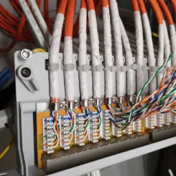 Panel krosowniczy z podłączonymi kablami sieciowymi, pomarańczowe kable zabezpieczone taśmą, widoczne złącza RJ45.