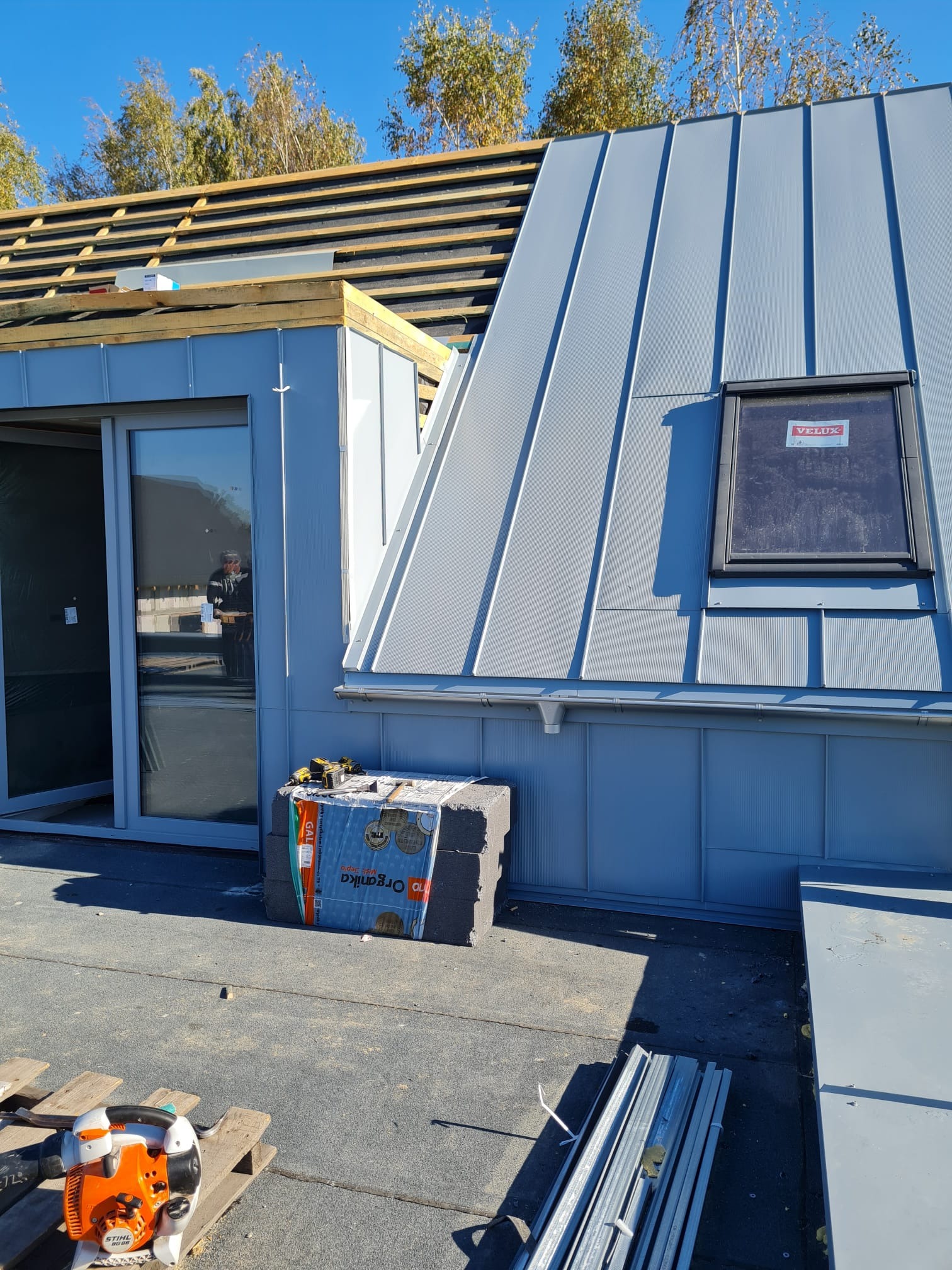 Szary dach z blachy na budynku z oknem dachowym Velux, widoczne narzędzia i materiały izolacyjne Organika, w tle drzewa.