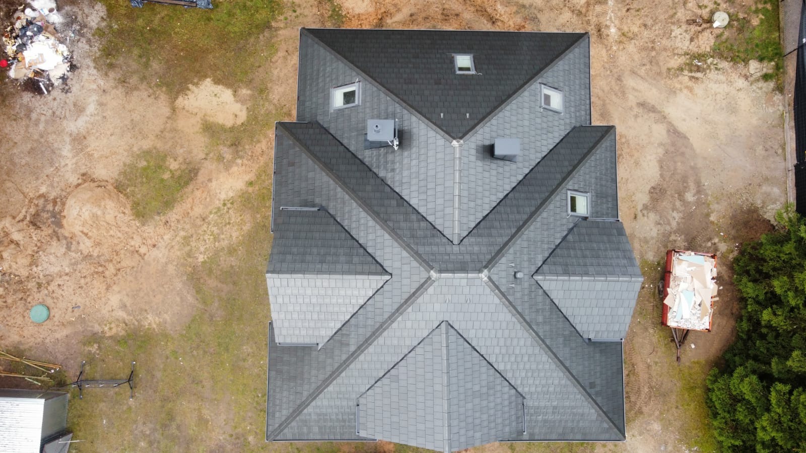 Widok z drona na dach domu z szarych dachówek bitumicznych o skomplikowanej geometrii, z kilkoma oknami dachowymi i kominami, obok kontener na odpady budowlane.