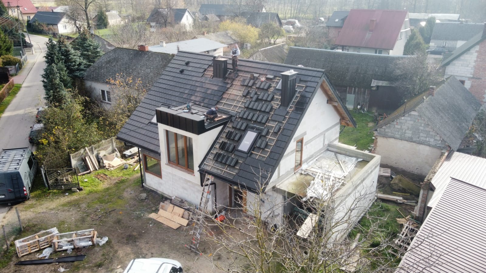 Montaż czarnej dachówki na dwuspadowym dachu z oknem połaciowym, widok z góry na budynek w trakcie prac dekarskich.