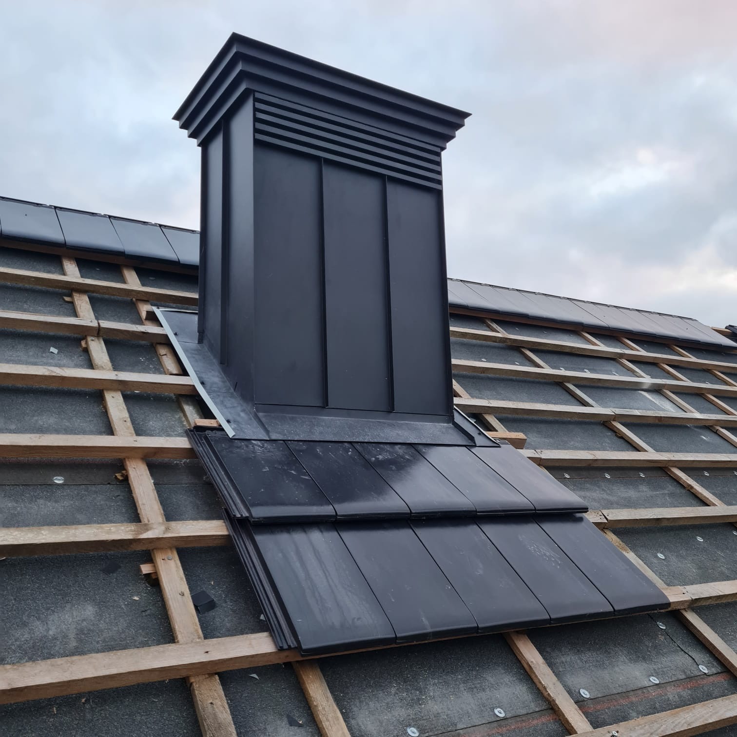 Montaż czarnego komina z blachy na dachu z dachówki ceramicznej w Warszawie, widoczne elementy konstrukcji drewnianej.