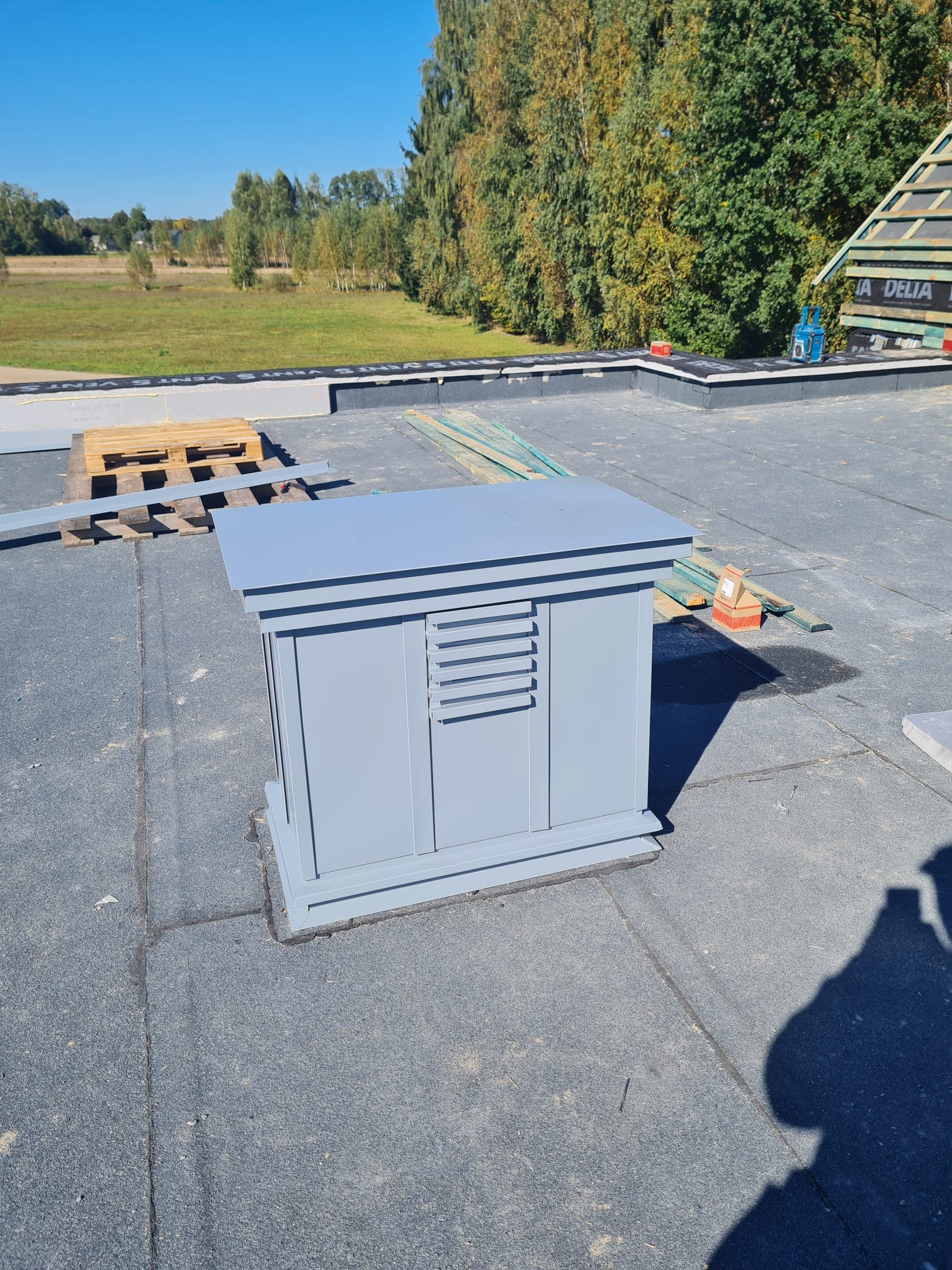 Szary element wentylacyjny na dachu pokrytym papą, w tle widoczne drzewa i fragment konstrukcji dachu z membraną DELTA.