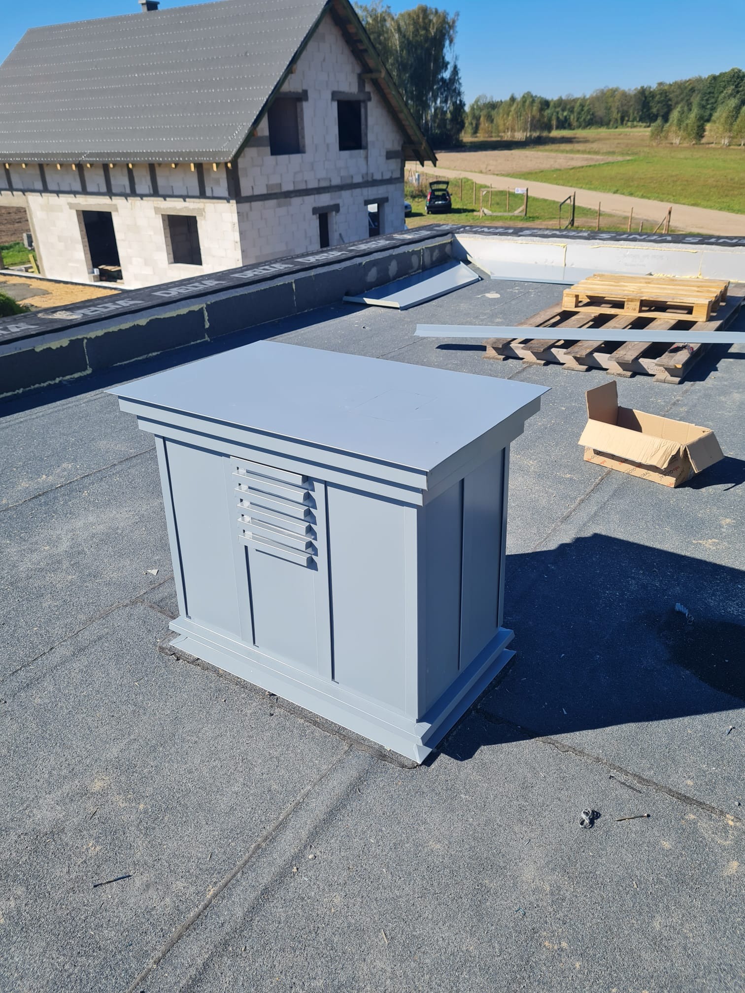 Szary kominek wentylacyjny na dachu pokrytym papą, w tle budynek w stanie surowym otwartym i palety z materiałami budowlanymi.