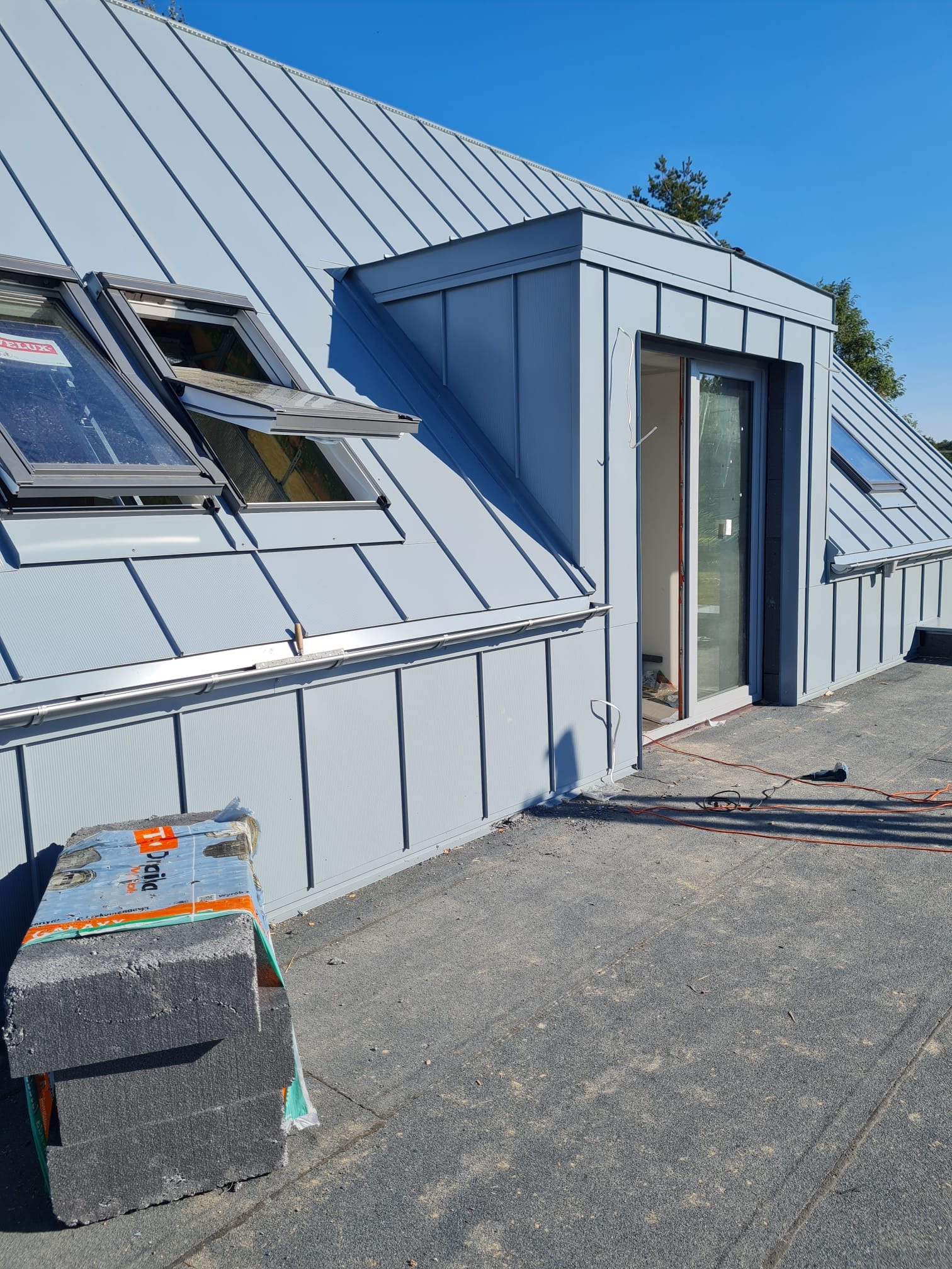 Nowoczesny, blaszany dach w kolorze gołębim z oknami dachowymi i wyjściem na dach, widok z góry na materiały budowlane i narzędzia.