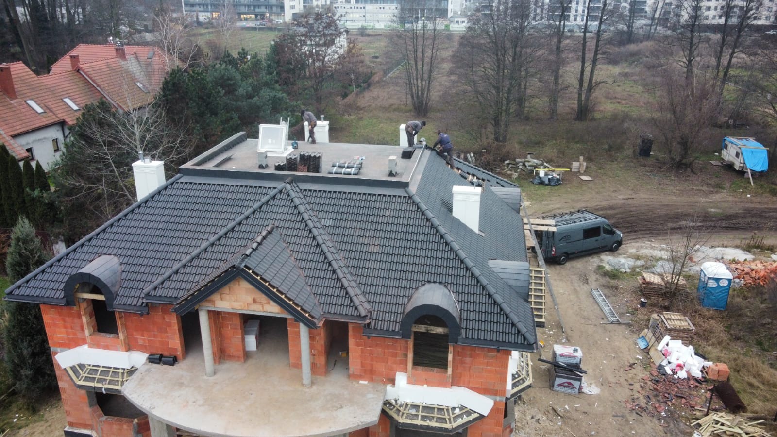 Ekipa dekarzy pracuje na dachu domu w stanie surowym, widoczna nowa, czarna dachówka, kominy, oraz samochód dostawczy na placu budowy.
