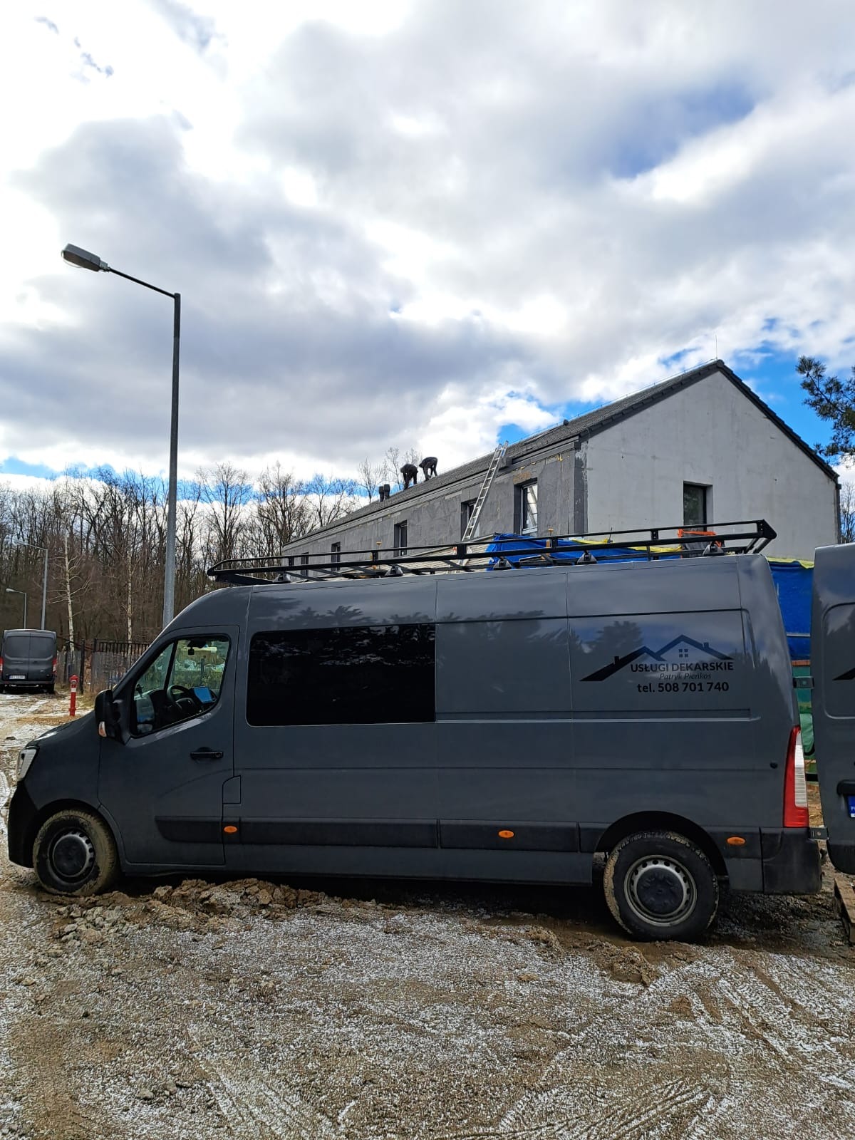 Szary samochód dostawczy z logo firmy dekarskiej na tle budynku z dachem w trakcie prac, na którym widać dwóch pracowników i drabinę.