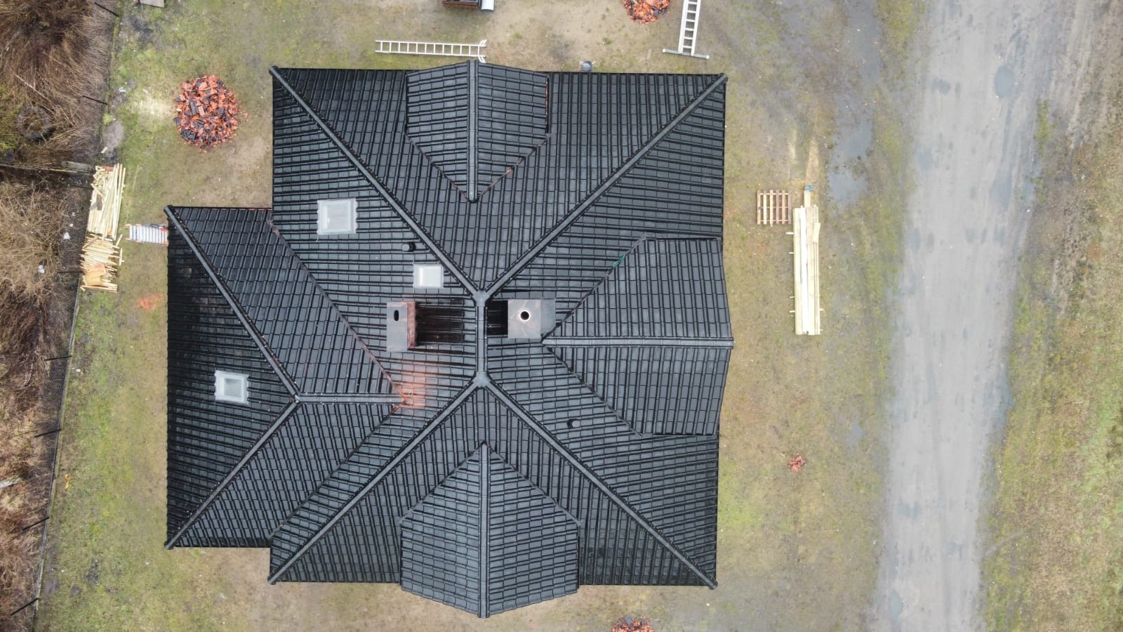 Widok z drona na czarny dach wielospadowy z lukarnami i kominami, obok sterty dachówek i przygotowane drewno na działce.