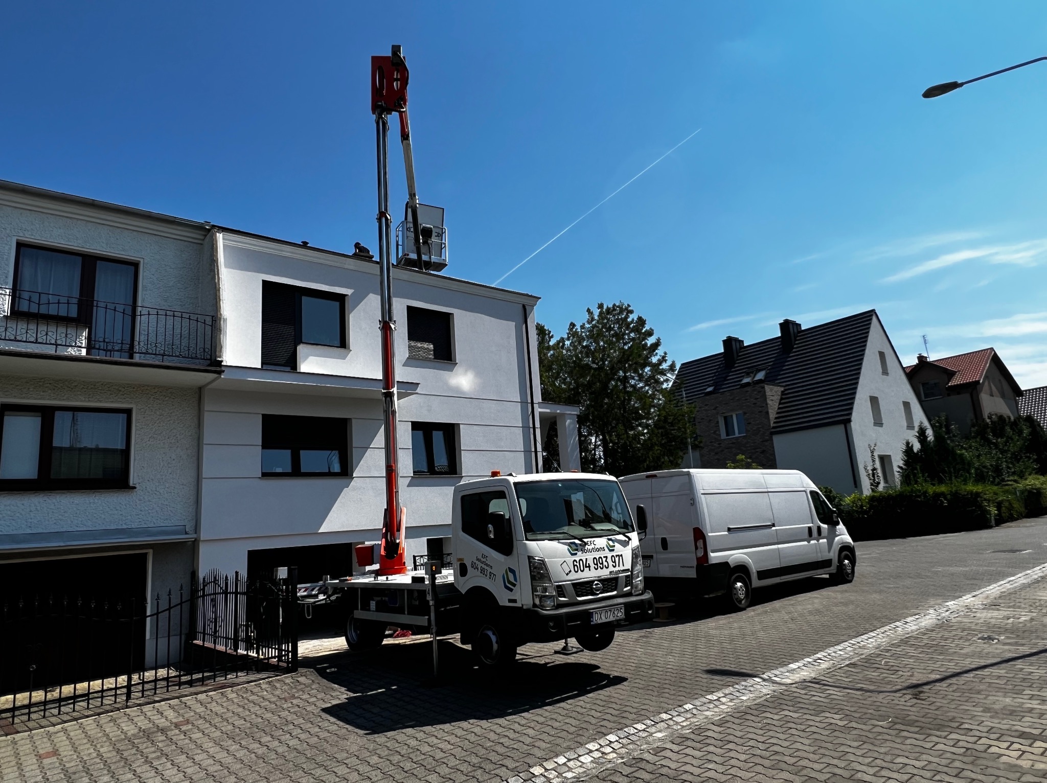 Pracownik w koszu podnośnika na wysokości dachu dwupiętrowego domu, biała elewacja, na chodniku stoi biała ciężarówka z podnośnikiem i biały bus, błękitne niebo z śladem kondensacyjnym samolotu.