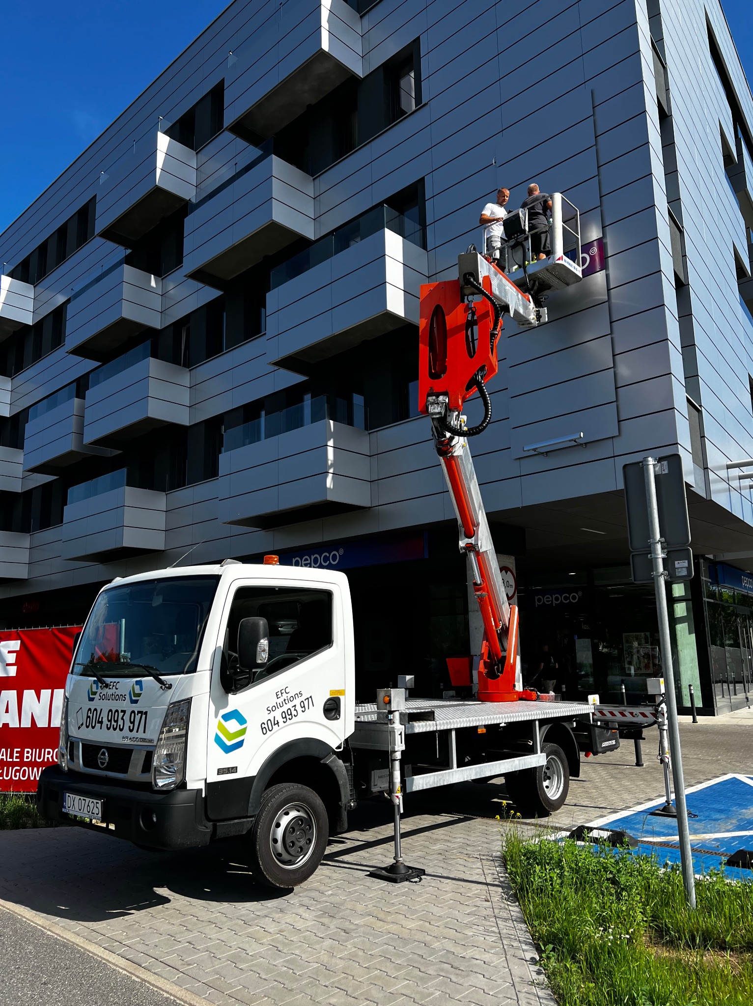 Dwaj pracownicy na platformie podnośnika koszowego, montują logo na elewacji nowoczesnego budynku biurowego, widoczny biały samochód dostawczy z logo firmy i numerem telefonu.