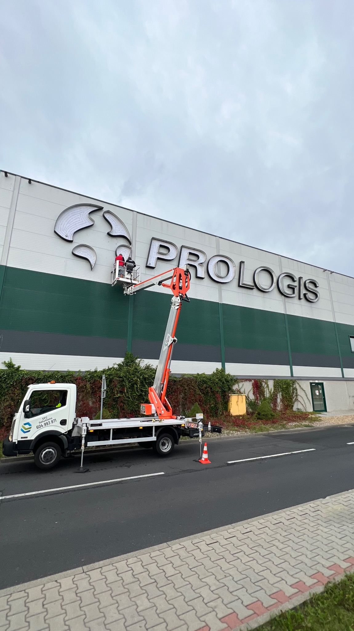 Montaż podświetlanego logo 'PROLOGIS' na elewacji budynku z użyciem podnośnika koszowego; widoczny pracownik w koszu i zaparkowany pojazd z podnośnikiem na tle zieleni.