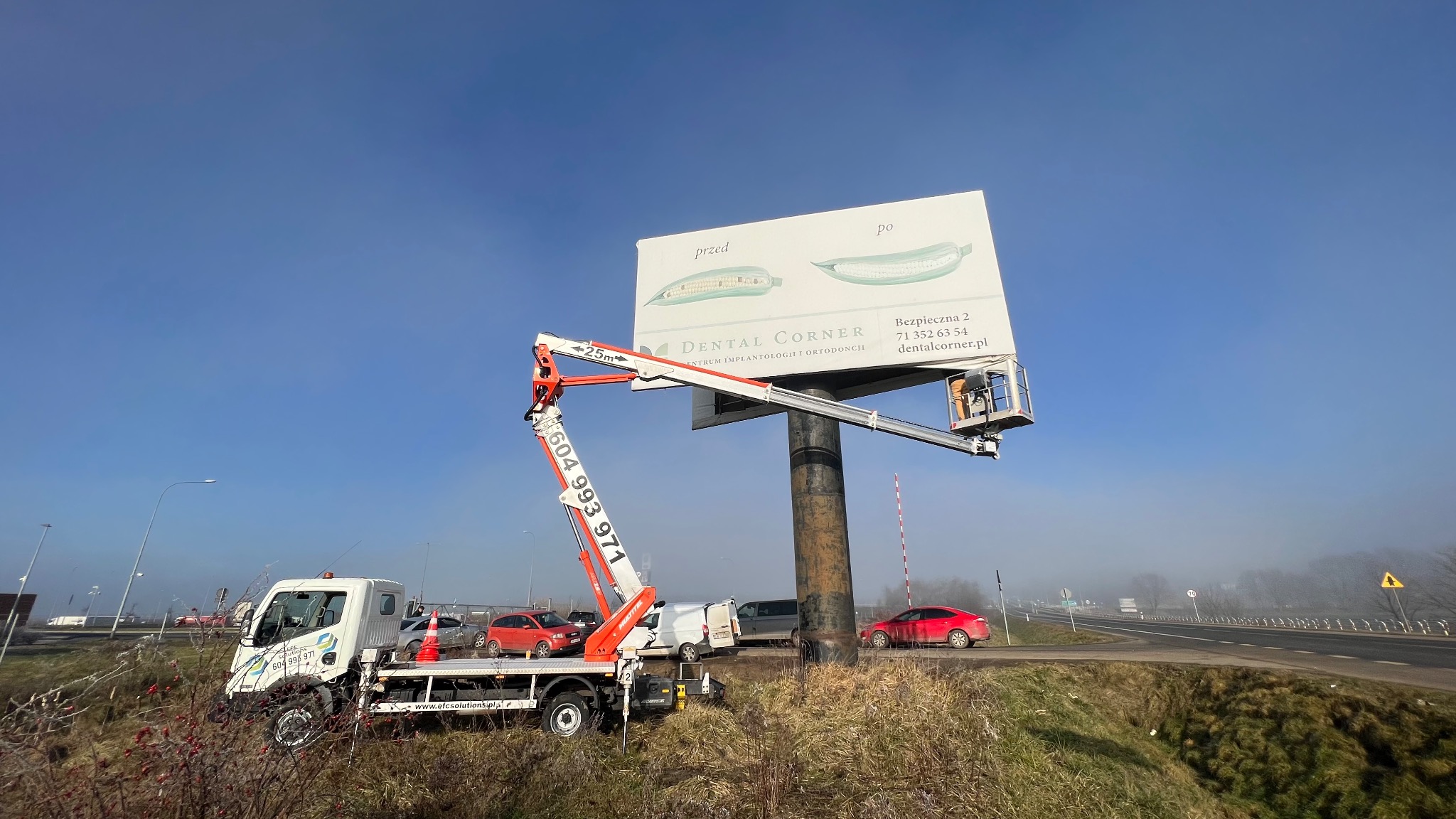 Montaż billboardu reklamowego z użyciem podnośnika koszowego; widoczny pracownik w koszu na tle błękitnego nieba i billboardu z reklamą dentystyczną.