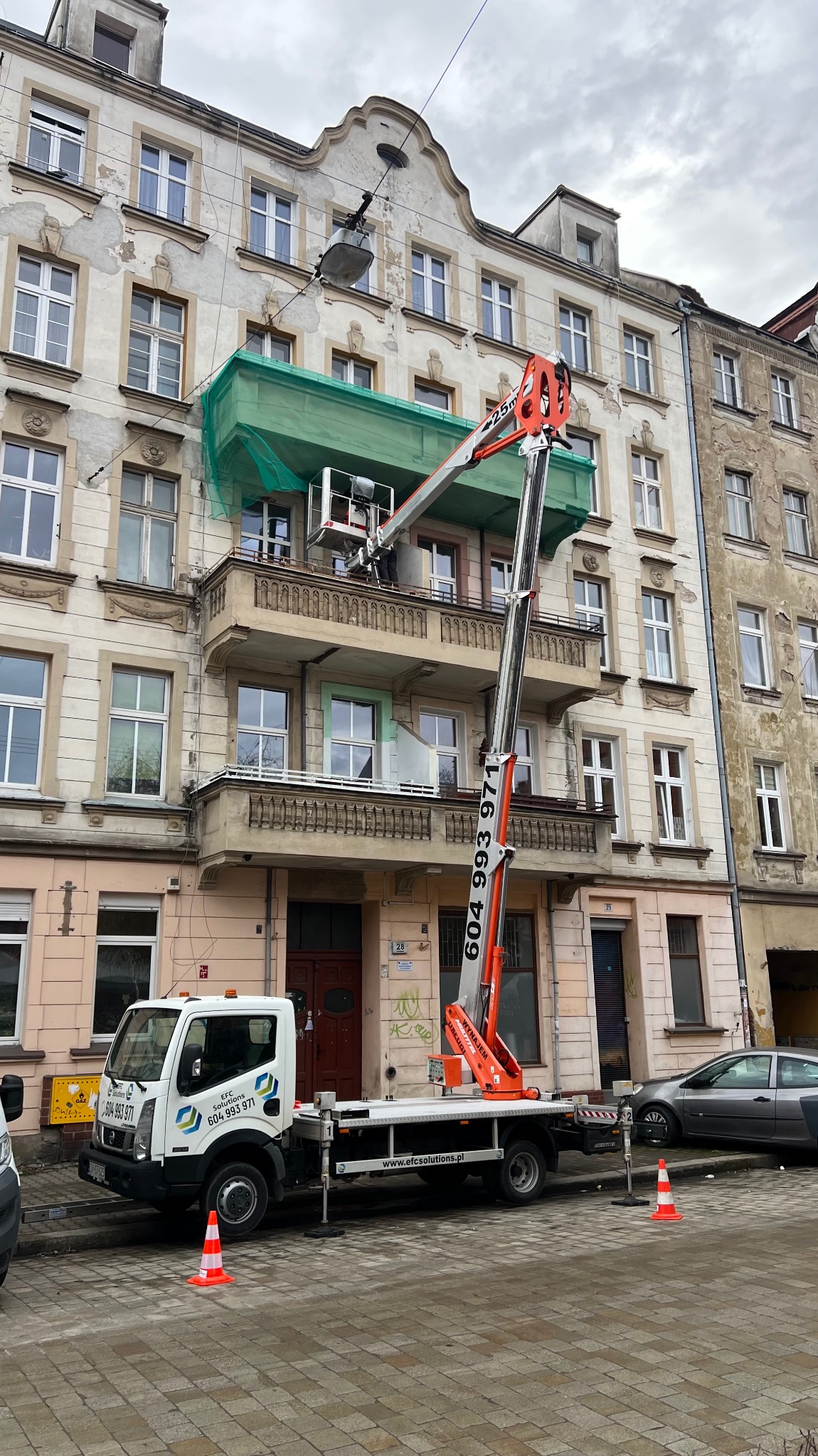 Prace na wysokości z wykorzystaniem podnośnika koszowego przy odnawianej elewacji budynku z balkonami, zabezpieczonej siatką ochronną