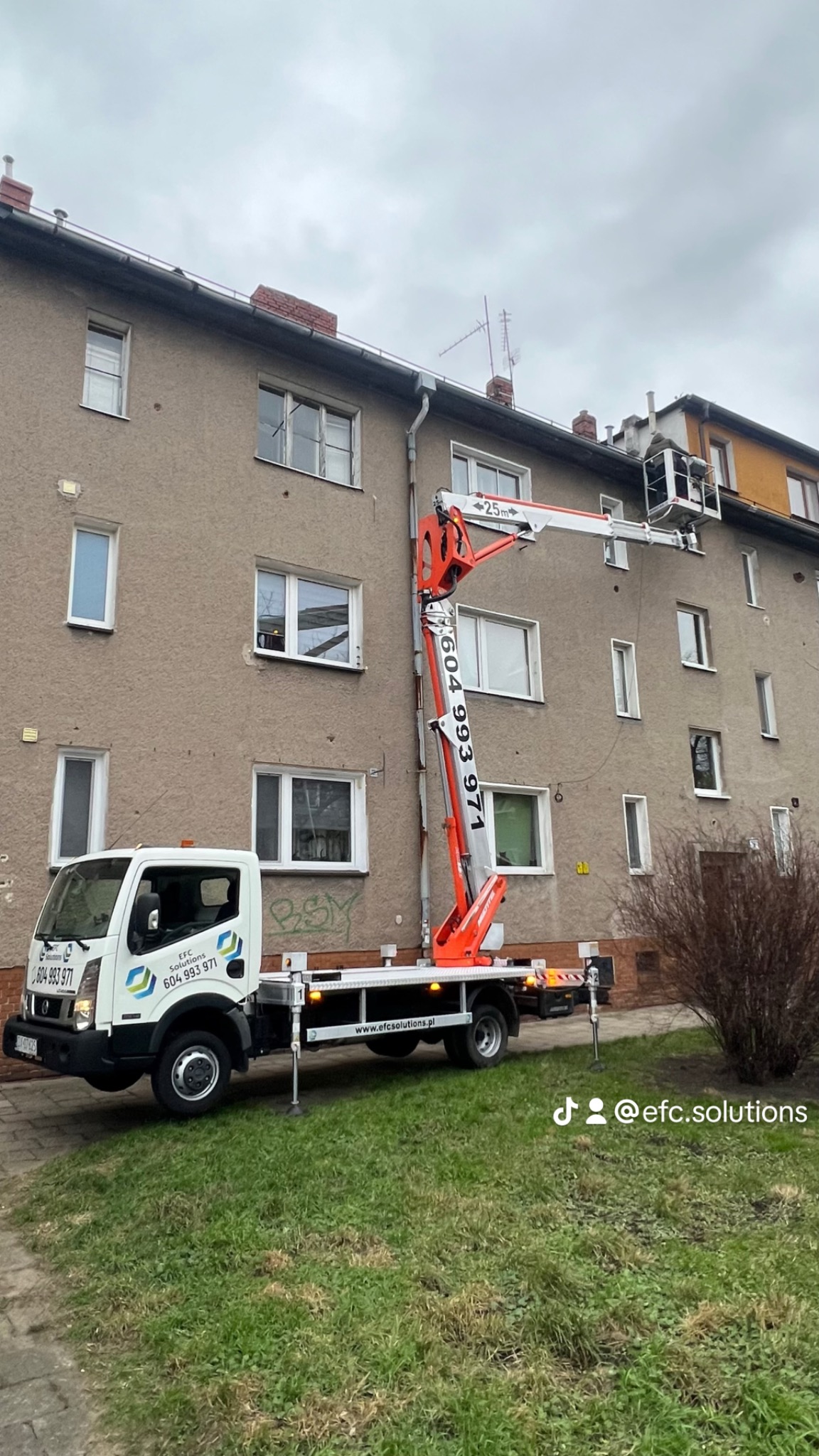 Pomarańczowo-biały podnośnik koszowy na tle bloku mieszkalnego z widocznym pracownikiem w koszu, przygotowującym się do pracy na wysokości.