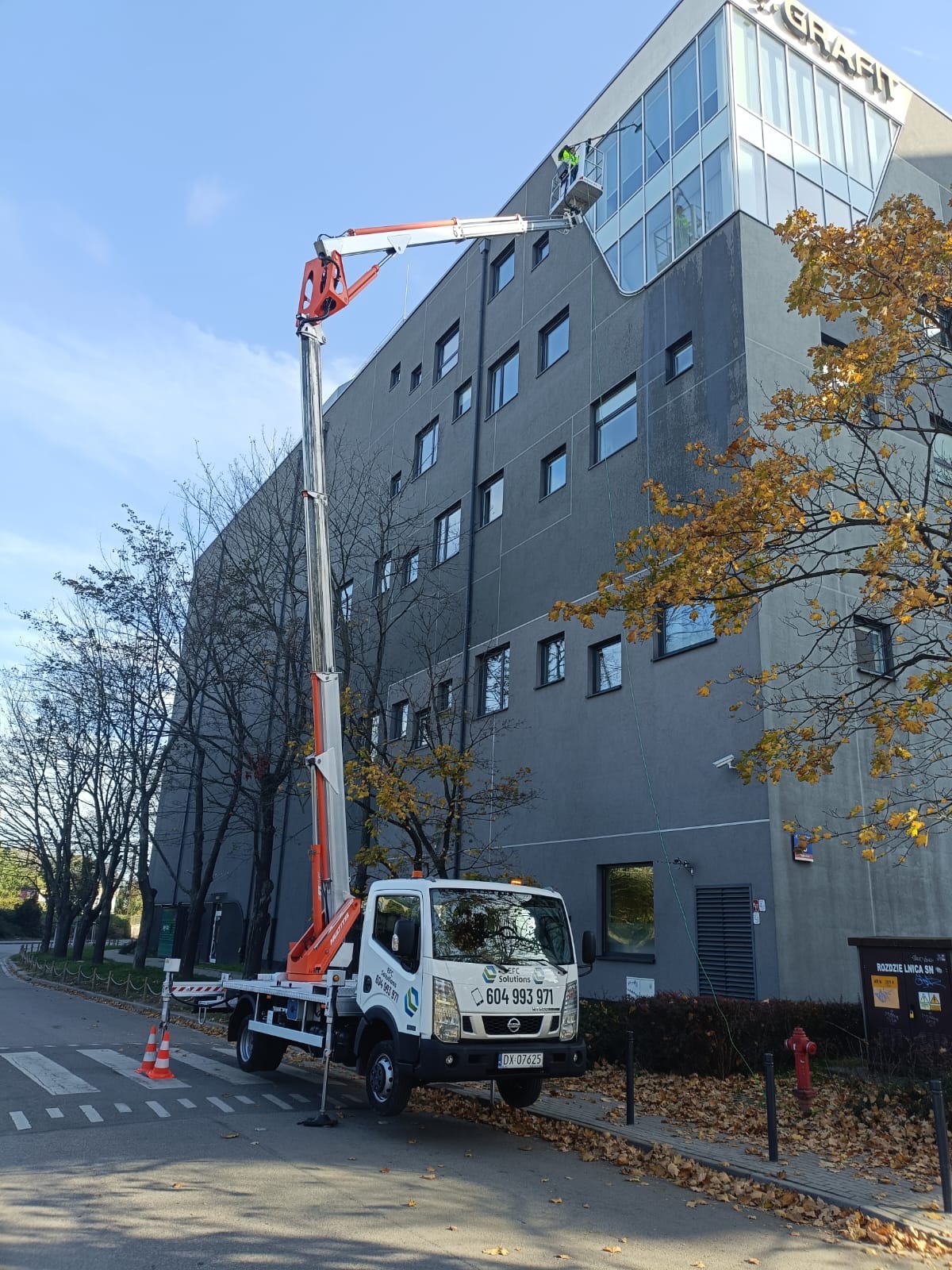 Pracownik w koszu podnośnika czyści okna na wysokości na elewacji biurowca, widoczny samochód z podnośnikiem oraz jesienne drzewa.