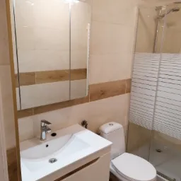 Jasna łazienka po remoncie: biała umywalka z szafką, toaleta i kabina prysznicowa, beżowe płytki z drewnopodobnym pasem, lustro z oświetleniem.