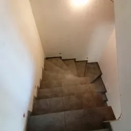 Wykończone schody wewnętrzne wyłożone płytkami ceramicznymi w odcieniach szarości, widok z góry na dół, z widocznymi śladami po wierceniu w ścianie.