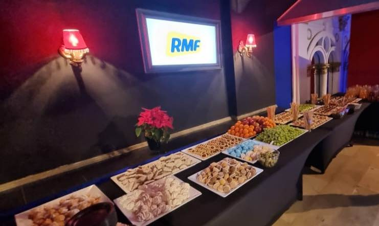 Stół z różnorodnymi przekąskami i owocami na czarnym obrusie, w tle ściana z dekoracyjnymi lampami, telewizor z logo RMF i ozdobna konstrukcja architektoniczna.