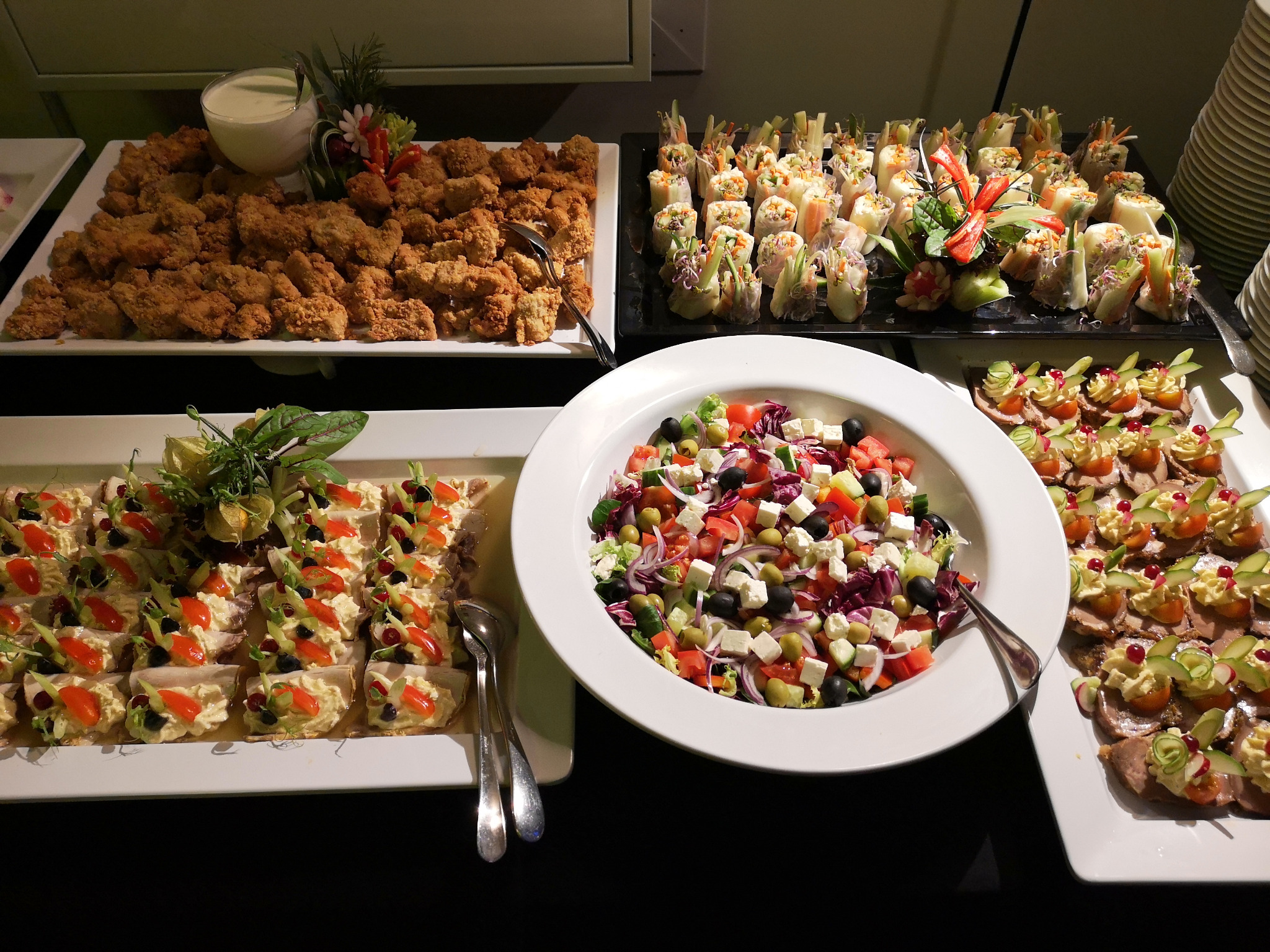 Bogato zastawiony stół z różnorodnymi przekąskami: smażone kąski z sosem, wiosenne roladki, sałatka grecka i kanapeczki z pastą, pomidorkami i ogórkiem.